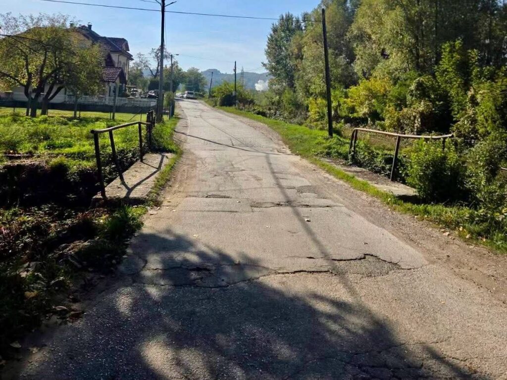 Oštećena asfaltna cesta s pukotinama i zakrpama između kuća i zelenila