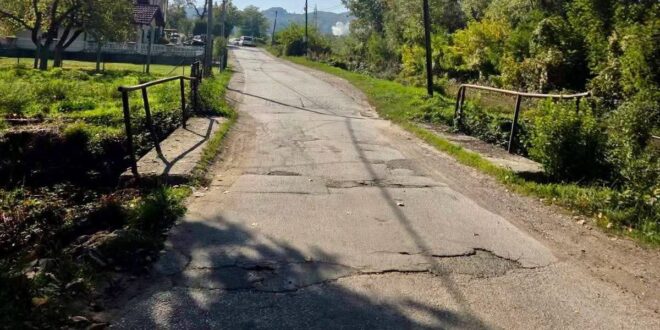 Oštećena asfaltna cesta s pukotinama i zakrpama između kuća i zelenila